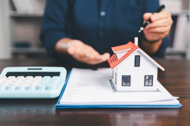 Masanın yanına yerleştirilen ev modeli madeni paraların yanı sıra, evrak işleri de gayrimenkul yatırımını, emlak değerini, mali planlama, mortgage seçeneklerini, emlak piyasasındaki pazar analizini simgeler..