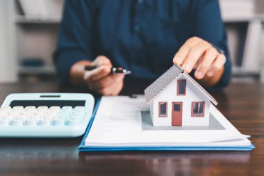 Masanın yanına yerleştirilen ev modeli madeni paraların yanı sıra, evrak işleri de gayrimenkul yatırımını, emlak değerini, mali planlama, mortgage seçeneklerini, emlak piyasasındaki pazar analizini simgeler..