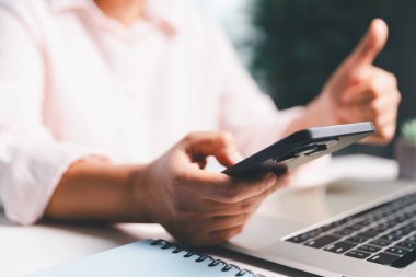 Yakın plan çekimde, kadın akıllı telefonunu kullanarak ahşap bir masada oturuyor, mobil bankacılık, online alışveriş ve sosyal medya ağlarında gezinirken parmağıyla telefon ekranına dokunuyor..