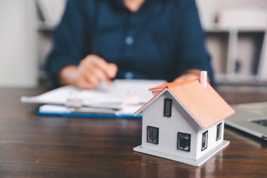 Masanın yanına yerleştirilen ev modeli madeni paraların yanı sıra, evrak işleri de gayrimenkul yatırımını, emlak değerini, mali planlama, mortgage seçeneklerini, emlak piyasasındaki pazar analizini simgeler..