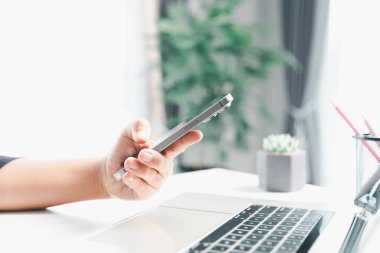 Yakın çekim sahnesinde, masada oturan bir kadın akıllı bir telefon tutuyor, modern e-randevu hizmetleri ve sosyal medyada mobil bir uygulama ile vakit geçiriyor, dijital kullanım kablosuz kavramını gösteriyor.