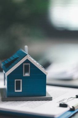 Emlakçı ve müşteri ev satın alma sözleşmesini tartışırken, arka planda emlak, sigorta ve kredi düzenlemelerini sembolize eden ev modeli süreçte yer alıyor.