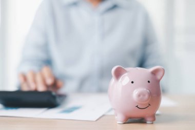 Tasarruf ve finansal güvenlik kavramı. Pembe kumbaralar ofis masasının üzerinde madeni paralarla çevrili. Arka planda iş kadını, yatırımı, zenginlik büyümesini ve bütçe yönetimini sembolize ediyor..