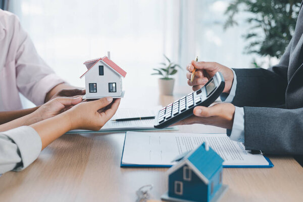 Close up of real estate agent handing house model to client with calculator and contract paperwork on desk, concept of mortgage loan, home ownership, property investment and housing agreement.