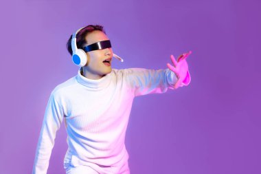 Beyaz kollu tişörtlü, VR gözlüklü ve kulaklık takan Asyalı oyuncu mor arka planda çalıp dokunuyor. Metaverse konsepti.