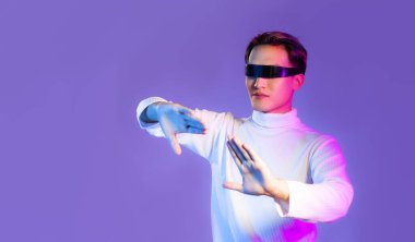 Beyaz, uzun kollu tişörtlü, VR gözlüklü, mor arka planda seyreden ve dokunan Asyalı bir genç. Metaverse konsepti.