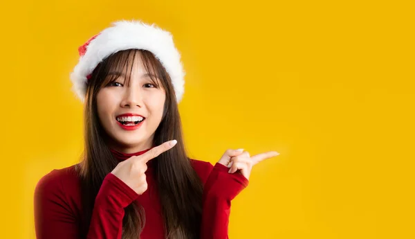 メリークリスマス 若いですかなりアジアの女性長いです髪型で赤長袖Tシャツ着用サンタ帽子指指す黄色の背景 — ストック写真