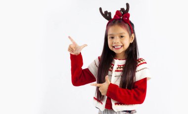 Kırmızı kazaklı küçük Asyalı çocuk Noel temalı kostümlü gülümseyen parmağıyla beyaz arka planı işaret ediyor. Mutlu Noeller..
