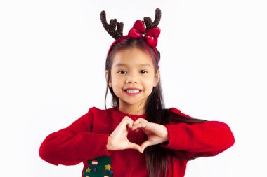 Kırmızı kazaklı küçük Asyalı çocuk Noel temalı kostümlü gülümsüyor ve beyaz arka planda kalp işareti yapıyor..