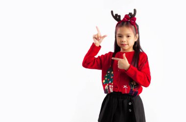 Kırmızı kazaklı küçük Asyalı çocuk Noel temalı kostüm giyip beyaz arka planda boş bir yeri işaret ediyor..
