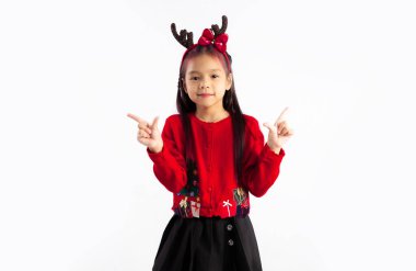 Kırmızı kazaklı küçük Asyalı çocuk Noel temalı kostüm giymiş kameraya bakıp beyaz arka planı işaret ediyor..