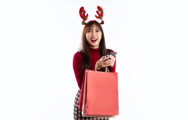 スマートフォンを使ってトナカイの角の帽子をかぶった赤いクリスマスの衣装を着た若いアジアの女性とショッピングバッグ白の背景を運ぶ メリークリスマス — ストック写真