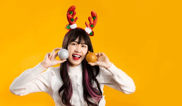 Pretty asian woman long hairstyle in white sweater wearing reindeer horns headband holding silver and gold glitter balls smiling and looking camera yellow background. Merry Christmas concept.