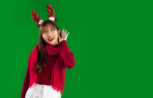 メリークリスマス 若いですかなりアジアの女性で赤と白のクリスマスのテーマの衣装を身に着けているトナカイ角ヘッドバンドポージングリスニングとゴシップ緑の画面の背景に — ストック写真
