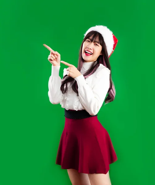 身穿红白相间圣诞主题服装的年轻漂亮的亚洲女人戴着桑塔帽 手指头指向绿色背景 圣诞快乐 — 图库照片
