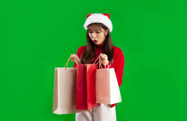 身穿红白相间的圣诞主题服装 头戴圣诞礼帽的年轻亚洲女人在打开购物袋绿色荧幕时大吃一惊 — 图库照片