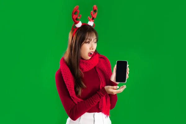 Asian woman long hairstyle in red sweater and scarf wearing reindeer horns headband holding and showing blank black screen smartphone posing surprised on green screen background