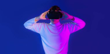 Beyaz uzun kollu tişörtlü Asyalı adam video gözlüklü kulaklık takıyor ve internette oyun oynarken koyu mavi arka plan izliyor. Metaverse konsepti