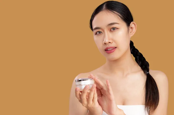 Skin care and beauty. Young asian woman clean face holding and showing a jar of cream skin care on beige background.