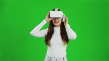 Front view of a young asian woman looking around, wearing a white shirt and a VR headset, greenscreen