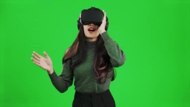 Virtual Reality concept. Front view of a young asian woman looking around, wearing a gray shirt and a VR headset, greenscreen