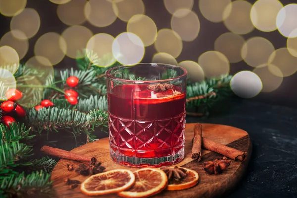 Mulled wine in a glass on wooden cutting board with spices, spruce branch and bokeh on a background, close up.