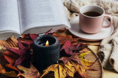 Sonbahar yapraklarıyla süslenmiş yanan mum, açık kitap ve kahve fincanı. Sıcak ev, sonbahar konsepti. Seçici odak.