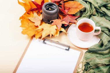 Boş panolu bir masaüstü, bir fincan çay, sonbahar yaprakları ve açık bej arka planda mum. Üst görünüm, model.
