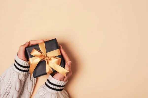 Girl\'s hands in white sweater holding black gift box with golden bow on neutral beige background. Christmas, Valentines day, Black friday sale concept. Top view, flat lay, copy space.