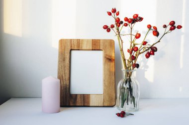 Beyaz masadaki boş resim çerçevesi, mum ve vazoda gül ağacı dalları. Ön görünüm, modelle.