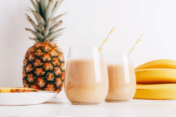 stock image Still life with fruits, banana, pineapple and smoothie in glasses on white wooden table. Minimal detox diet concept, summer vitamin drink. Front view.