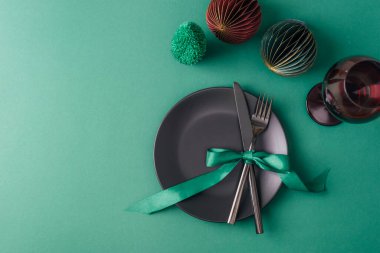 Siyah tabak, çatal ve bıçak, şarap kadehi ve yeşil arka planda Noel süslemeleri olan şenlik masası. Üst görünüm, düz görünüm, kopyalama alanı.