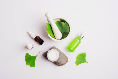 Taze yeşil ginkgo yaprakları, harç ve tokmak, beyaz arka planda kozmetik serumu şişesi ve krema kavanozu. Doğal el yapımı kozmetik konsepti. Üst manzara, düz uzanma.