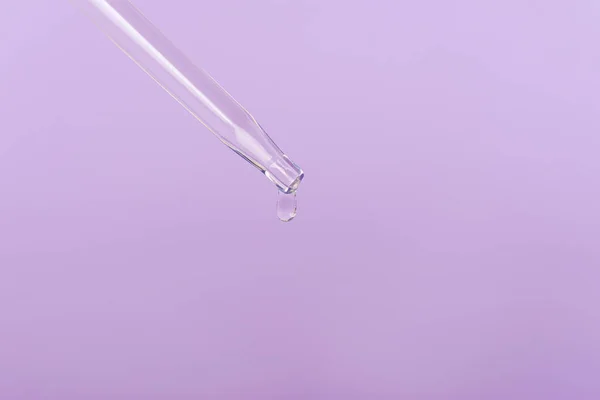 Drop of cosmetic face serum on a pipette on light purple background. Front view, closeup. Copy space.