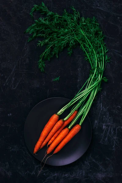 Ripe carrot with greens on black plate on black background. Healthy vegan food. Top view, flat lay.