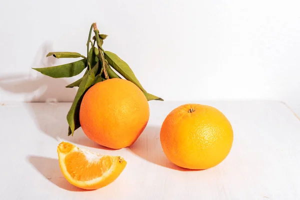 Fresh ripe oranges with leaves and orange slice on white table in sunlight. Healthy food concept, closeup.