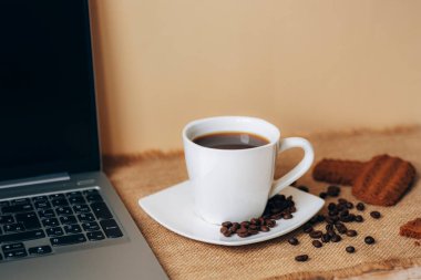 Coffee cup with beans, cookies and laptop on beige table.