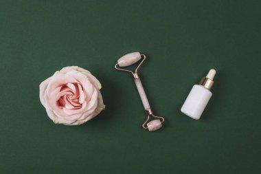 White cosmetic bottles, facial roller and pink rose flower on dark green background. Natural cosmetics, skin care concept. Top view, flat lay.
