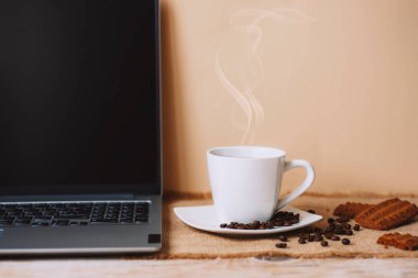 White cup of hot coffee with beans, cookies and laptop on beige table background.