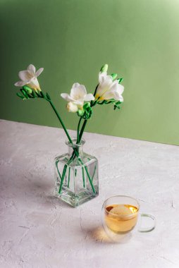 A cup of tea and freesia white flowers in glass vase on white textured table against green wall. Still life.