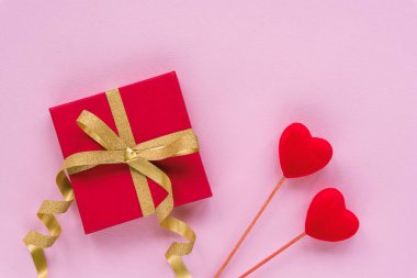 Gift in a red box with golden bow and two toy hearts on pink background. Valentines day concept. Top view, flat lay.