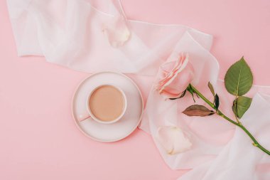 A coffee cup and pink rose on white silk fabric. Holiday concept. Top view, flat lay.