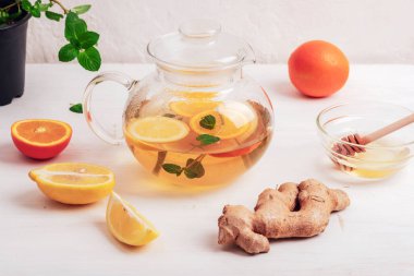 Glass teapot with orange, lemon, ginger and mint tea with ingredients on white wooden table.