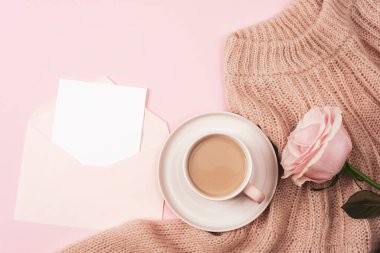 A coffee cup, pink rose on knitted sweater and envelope with blank card. Holiday concept. Top view, flat lay, mockup.
