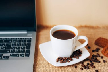 Black coffee in white cup with beans, cookies and laptop on a table. Top view, closeup.