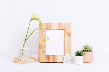 Mockup wooden picture frame, carved decor heart and freesia flower in vase on white table. Copy space.