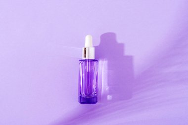 Purple cosmetic face serum bottle on palm leaf shadow background in sunlight. Top view, flat lay.