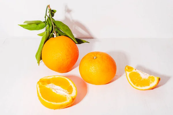 Fresh ripe oranges with leaves and orange slices on white table in sunlight. Healthy food concept, closeup.