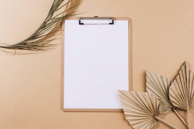 Desktop with blank clipboard, fan leaves and dried palm leaf on beige background. Top view, flat lay, mockup.