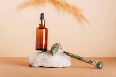 Brown cosmetic serum bottle and jade facial roller on a natural stone podium. Natural skin care cosmetics concept. Closeup, copy space.